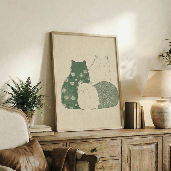May include: Framed artwork showing three cats in soft green and cream colors, with one cat featuring a white daisy pattern, displayed on a rustic wooden sideboard alongside books, a ceramic vase, and a fern plant in a cozy living room setting with neutral tones and natural lighting.