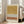 May include: A framed abstract art print leaning against a white wall next to a beige sofa in a bright living room. The artwork features horizontal bands of mustard yellow watercolor, coral orange brushstrokes, and black polka dot patterns on cream background. Natural sunlight streams through tall windows, casting soft shadows on light wooden floors.