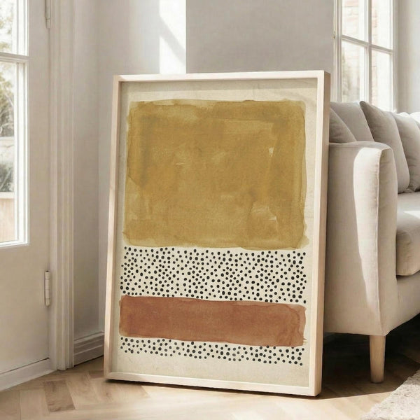 May include: A framed abstract art print leaning against a white wall next to a beige sofa in a bright living room. The artwork features horizontal bands of mustard yellow watercolor, coral orange brushstrokes, and black polka dot patterns on cream background. Natural sunlight streams through tall windows, casting soft shadows on light wooden floors.
