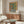 May include: Framed abstract cactus artwork displayed above wooden mid-century modern credenza in bright living room. Green saguaro cactus in black and white striped pot against warm orange gradient background. White curved chair and glass coffee table visible in foreground with natural lighting creating soft shadows across cream walls.