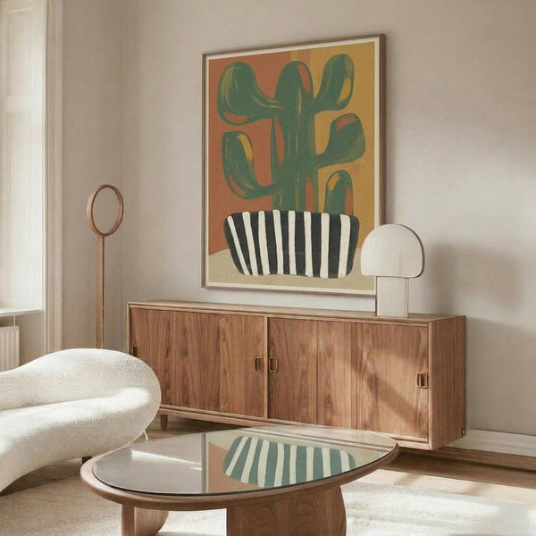 May include: Framed abstract cactus artwork displayed above wooden mid-century modern credenza in bright living room. Green saguaro cactus in black and white striped pot against warm orange gradient background. White curved chair and glass coffee table visible in foreground with natural lighting creating soft shadows across cream walls.