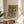 May include: Framed abstract cactus artwork displayed on a round wooden side table next to a beige upholstered chair. The poster features a green saguaro cactus in a black and white striped pot against a warm orange and yellow gradient background. Natural sunlight streams across the neutral-toned room setting with a plant visible in the corner.