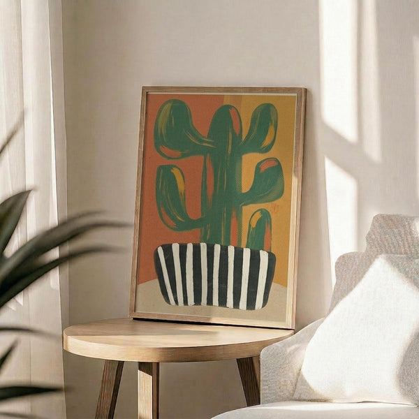 May include: Framed abstract cactus artwork displayed on a round wooden side table next to a beige upholstered chair. The poster features a green saguaro cactus in a black and white striped pot against a warm orange and yellow gradient background. Natural sunlight streams across the neutral-toned room setting with a plant visible in the corner.