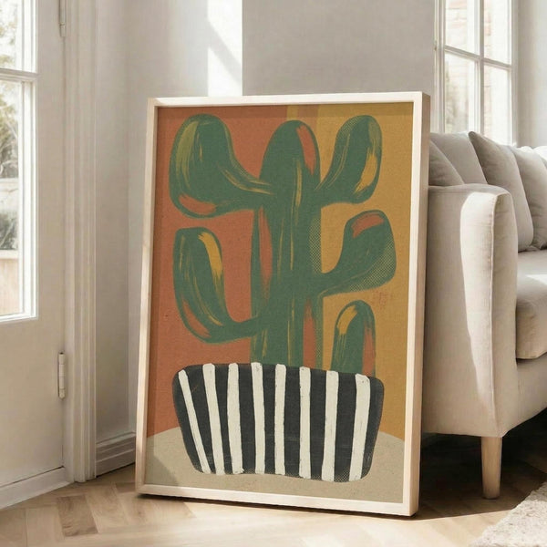 May include: A framed poster print featuring a green saguaro cactus in a black and white striped pot against a warm orange and yellow background. The artwork has a vintage, textured appearance and is displayed in a light wood frame leaning against a white wall next to a beige sectional sofa in a bright, modern living room with large windows.
