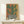 May include: Framed abstract cactus print in warm desert tones leaning against white wall on light wood surface, featuring green saguaro cactus in black and white striped pot with terracotta and golden yellow background, small white wrapped gift with greenery beside frame