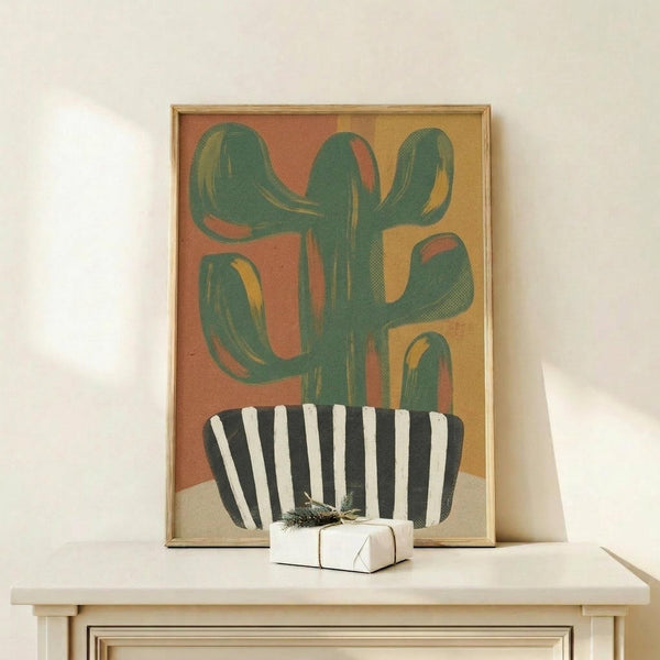May include: Framed abstract cactus print in warm desert tones leaning against white wall on light wood surface, featuring green saguaro cactus in black and white striped pot with terracotta and golden yellow background, small white wrapped gift with greenery beside frame