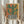 May include: Person holding framed abstract cactus artwork in light wood frame. Green saguaro cactus with curved arms against warm orange and yellow gradient background, planted in black and white striped pot. Modern minimalist style print displayed in bright interior with dried pampas grass and white curtains visible.