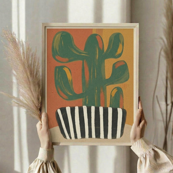 May include: Person holding framed abstract cactus artwork in light wood frame. Green saguaro cactus with curved arms against warm orange and yellow gradient background, planted in black and white striped pot. Modern minimalist style print displayed in bright interior with dried pampas grass and white curtains visible.