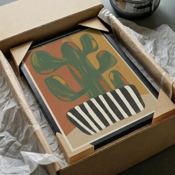 May include: Framed abstract art print featuring a stylized green cactus in a black and white striped pot against warm orange and yellow background, displayed in black frame inside open cardboard shipping box with white tissue paper and wooden packing materials on white surface