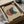 Abstract geometric artwork featuring two overlapping circles in terracotta orange and deep blue-green on a cream background, displayed in a black frame inside an open cardboard shipping box with protective tissue paper and wooden corner guards