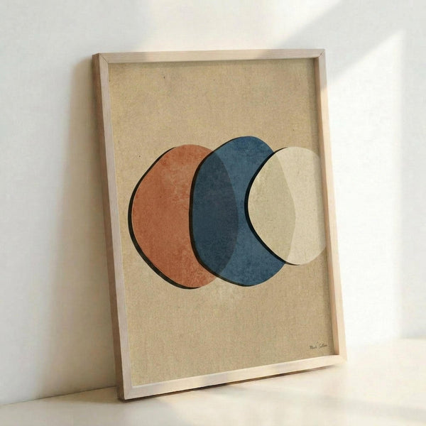 May include: Framed abstract art print featuring three overlapping circles in terracotta orange, deep blue, and cream colors on textured beige paper background. Light wood frame sits propped against white wall, casting soft shadows. Minimalist geometric design with organic, hand-painted texture visible in each circle.