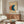 May include: Modern living room with mid-century walnut credenza displaying abstract geometric artwork featuring three overlapping circles in coral orange, teal blue, and cream on beige background in thin brown frame. White sculptural chair, glass coffee table, and minimalist white table lamp complete the styled interior scene.