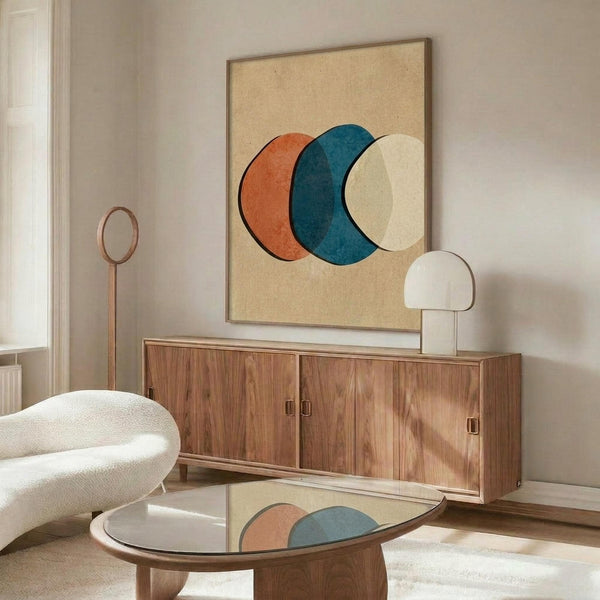 May include: Modern living room with mid-century walnut credenza displaying abstract geometric artwork featuring three overlapping circles in coral orange, teal blue, and cream on beige background in thin brown frame. White sculptural chair, glass coffee table, and minimalist white table lamp complete the styled interior scene.