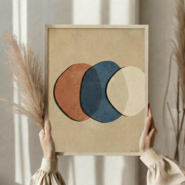 May include: Person in beige clothing holding framed abstract art print featuring three overlapping circles in terracotta orange, navy blue, and cream on textured beige background. Natural wood frame, dried pampas grass visible to left, soft neutral lighting and curtains in background.