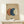 May include: Abstract art print with three overlapping circles in terracotta orange, navy blue, and cream on beige textured background, displayed in light wood frame on white wooden console table beside small wrapped gift with evergreen sprig against white wall