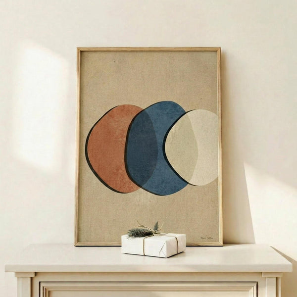 May include: Abstract art print with three overlapping circles in terracotta orange, navy blue, and cream on beige textured background, displayed in light wood frame on white wooden console table beside small wrapped gift with evergreen sprig against white wall