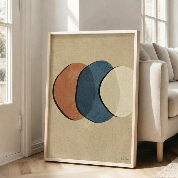 May include: Abstract art print in light wood frame leaning against white wall next to beige sofa. Artwork features three overlapping circles in rust orange, navy blue, and cream on textured beige background. Modern minimalist living room setting with natural light from windows, clean white walls and neutral furniture.