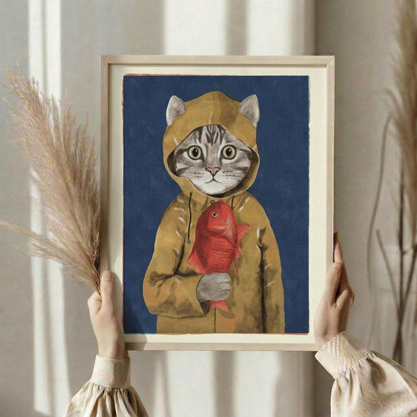 May include: Person holding framed artwork showing a tabby cat wearing a mustard yellow hooded raincoat and holding a red fish, set against a navy blue background. The wooden frame has a light natural finish with white matting. Dried wheat stalks are visible in the left hand, and the scene is photographed against white curtains in soft natural lighting.
