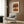 May include: Abstract artwork with horizontal brushstrokes in terracotta, ochre, and brown tones featuring dotted stipple patterns down the center, displayed in light wood frame on textured wood wall above modern cream armchair with geometric pillow, alongside brass floor lamp and dark side table with open book.