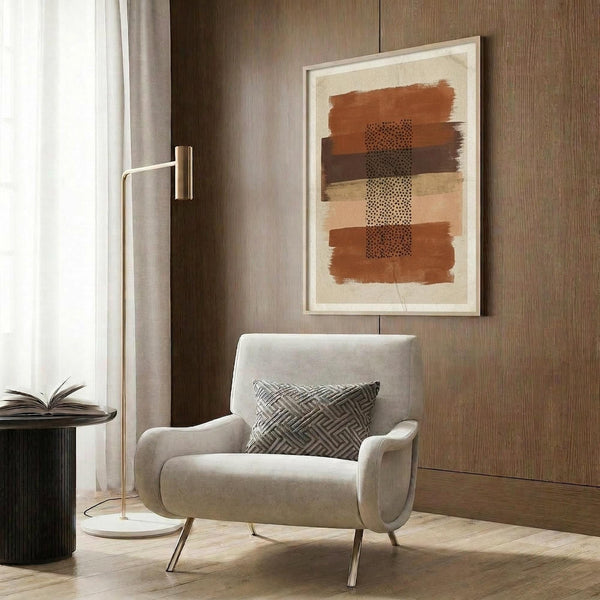 May include: Abstract artwork with horizontal brushstrokes in terracotta, ochre, and brown tones featuring dotted stipple patterns down the center, displayed in light wood frame on textured wood wall above modern cream armchair with geometric pillow, alongside brass floor lamp and dark side table with open book.