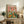 May include: Framed poster with colorful illustration of two white ducks in Hawaiian shirts and hats at picnic table with bread, cheese, wine bottles and rubber ducklings in basket, displayed on rustic wooden console with books, plant and pottery in neutral living room setting