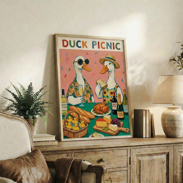 May include: Framed poster with colorful illustration of two white ducks in Hawaiian shirts and hats at picnic table with bread, cheese, wine bottles and rubber ducklings in basket, displayed on rustic wooden console with books, plant and pottery in neutral living room setting