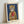 May include: Framed art print leaning against white wall showing tabby cat wearing yellow hooded raincoat holding red fish, illustrated in realistic style against deep blue background, displayed in light wood frame with white mat