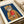 May include: Framed art print showing a tabby cat wearing a yellow hooded raincoat and holding a red fish, set against a deep blue background. The illustration has a whimsical, hand-painted style with visible brushstrokes. The print sits in a light wooden frame with white matting on a cream-colored surface next to rolled paper and ribbon.
