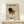 May include: Framed Kandinsky-style abstract geometric poster featuring black circle, red triangles, yellow circle, blue square, and diagonal black lines on cream background. Wooden frame displayed on light wood shelf next to small wrapped gift with twine. Clean white wall background.