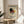 May include: Framed Kandinsky-style abstract art poster with geometric shapes including black circle, red triangles, yellow circle, and intersecting black lines on cream background, displayed on round wooden side table next to beige upholstered chair with decorative pillow and potted plant in bright, sunlit room with white walls.