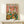 May include: Framed poster print leaning against white wall on light wooden shelf, featuring two cartoon ducks in colorful shirts at picnic table with wine bottles, cheese, bread, and basket of rubber duckies. Pink confetti background, "DUCK PICNIC" title in rainbow letters. Small white wrapped gift sits in front of natural wood frame.