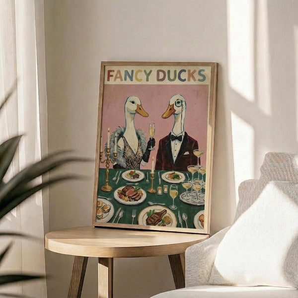 May include: Framed vintage-style poster showing two anthropomorphic ducks in formal evening wear dining at an elegant table. The female duck wears a sequined dress with feathers, the male a burgundy tuxedo with bow tie. They're seated at a green tablecloth-covered table laden with fine dining plates, wine glasses, candles, and gourmet food. The artwork has a pink background with "FANCY DUCKS" in colorful lettering at the top. The wooden-framed print sits on a round wooden side table next to a