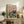 May include: Vintage-style poster featuring two elegant ducks in formal attire dining at a green tablecloth-covered table with wine glasses, candles, and plated meals against a pink background, displayed in a light wood frame on a rustic wooden sideboard alongside books, a ceramic vase, and fern plant in a cozy living room setting with beige upholstered furniture.