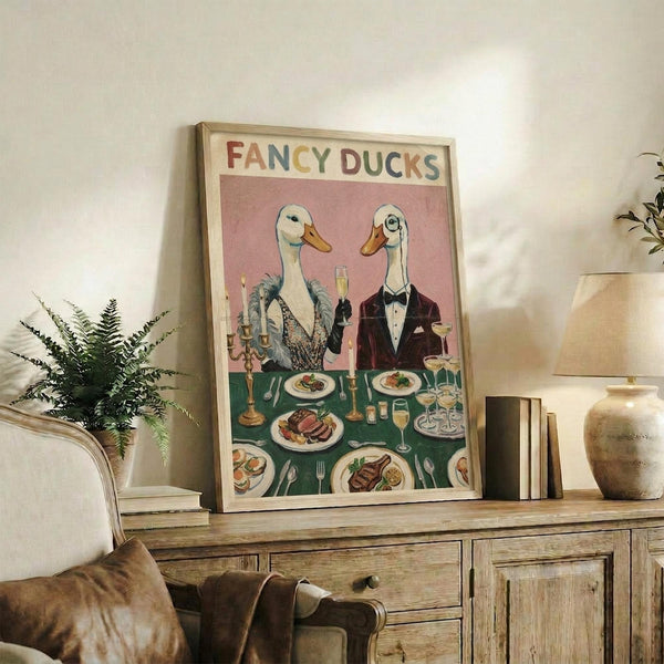 May include: Vintage-style poster featuring two elegant ducks in formal attire dining at a green tablecloth-covered table with wine glasses, candles, and plated meals against a pink background, displayed in a light wood frame on a rustic wooden sideboard alongside books, a ceramic vase, and fern plant in a cozy living room setting with beige upholstered furniture.