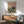 May include: Vintage-style poster showing two elegant ducks in formal evening wear seated at a dining table with candles, wine glasses, and plated meals. The illustration features warm pink and green tones with "Fancy Ducks" text at top. Displayed in wooden frame above mid-century modern credenza with white ceramic lamp, alongside sage green upholstered armchair in bright, naturally-lit room.