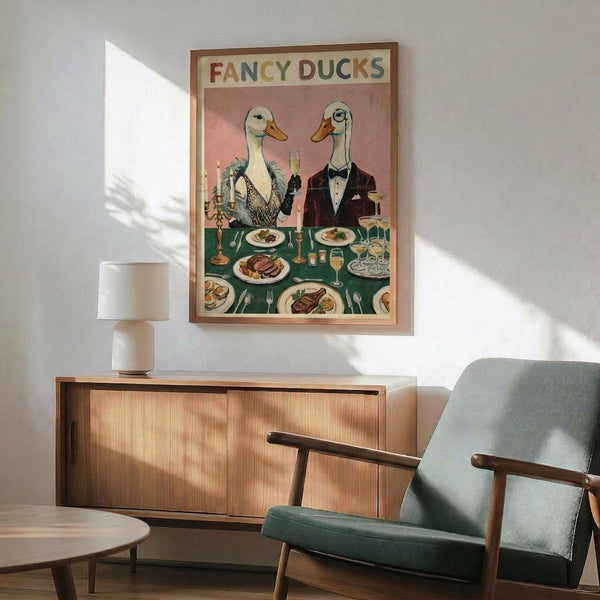May include: Vintage-style poster showing two elegant ducks in formal evening wear seated at a dining table with candles, wine glasses, and plated meals. The illustration features warm pink and green tones with "Fancy Ducks" text at top. Displayed in wooden frame above mid-century modern credenza with white ceramic lamp, alongside sage green upholstered armchair in bright, naturally-lit room.