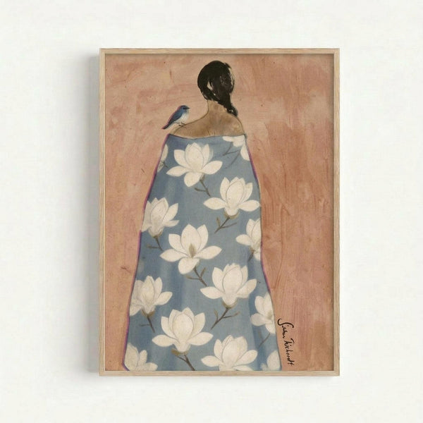Framed artwork showing a woman viewed from behind with dark hair in a braid, draped in a blue robe decorated with white magnolia flowers. A small blue bird perches on her bare shoulder. The background has a soft peachy-pink tone with subtle texture. The piece is displayed in a light wood frame against a white wall, with an artist signature visible in the bottom right corner.