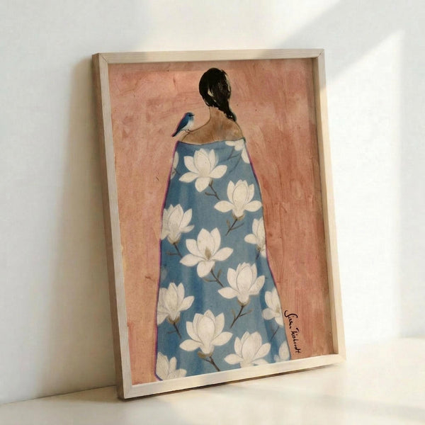 May include: Framed art print leaning against white wall showing woman from behind wearing blue floral robe with white magnolia blossoms, dark hair in bun with small blue bird perched on shoulder, painted on peachy pink background, displayed in light wooden frame on white surface
