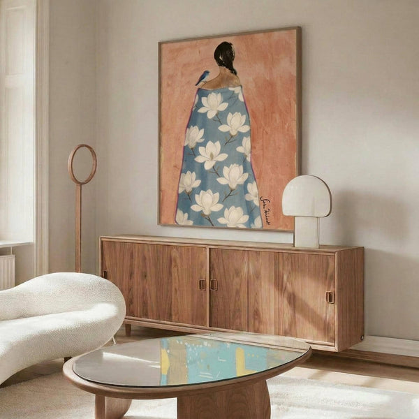 May include: Framed artwork showing a woman from behind wearing a blue floral robe with white magnolia blooms against a coral background, displayed above a mid-century modern walnut credenza in a minimalist living room with white upholstered chair, brass floor lamp, oval glass coffee table, and natural lighting creating warm shadows on neutral walls.