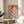 May include: Framed art print displayed on round wooden side table next to beige upholstered chair and potted plant. Artwork shows woman from behind wearing blue robe with white magnolia flower pattern against peachy pink background. Natural sunlight creates geometric shadows on white wall. Print has thin wooden frame and appears medium-sized relative to furniture.