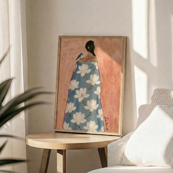 May include: Framed art print displayed on round wooden side table next to beige upholstered chair and potted plant. Artwork shows woman from behind wearing blue robe with white magnolia flower pattern against peachy pink background. Natural sunlight creates geometric shadows on white wall. Print has thin wooden frame and appears medium-sized relative to furniture.