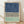 May include: Framed abstract art print leaning against white wall on light wood floor. Features three horizontal sections - textured navy blue at top, black and white herringbone chevron pattern in middle, and soft sage green at bottom. Natural wood frame with white matting. Sunlight creates diagonal shadows across the white wall.