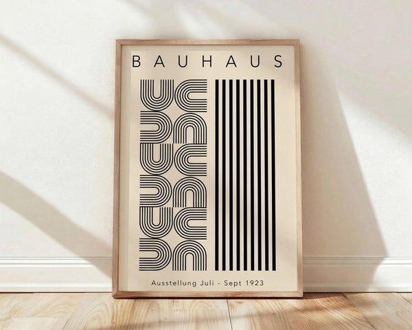 May include: Framed Bauhaus exhibition poster in light wood frame leaning against white wall on light wooden floor. Design features geometric black line patterns forming repeated semicircles and curves alongside bold vertical stripes on cream background. Clean, minimalist Scandinavian-style room setting with soft natural lighting and white fabric visible in background.
