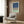 May include: Modern beige armchair with geometric throw pillow positioned beside tall brass floor lamp in contemporary room setting. Framed abstract artwork with deep blue, sage green, and herringbone pattern sections hangs on wood-paneled wall. Dark round side table with open book and white curtains complete the styled living space scene.