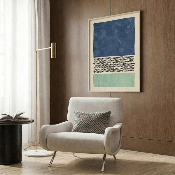 May include: Modern beige armchair with geometric throw pillow positioned beside tall brass floor lamp in contemporary room setting. Framed abstract artwork with deep blue, sage green, and herringbone pattern sections hangs on wood-paneled wall. Dark round side table with open book and white curtains complete the styled living space scene.