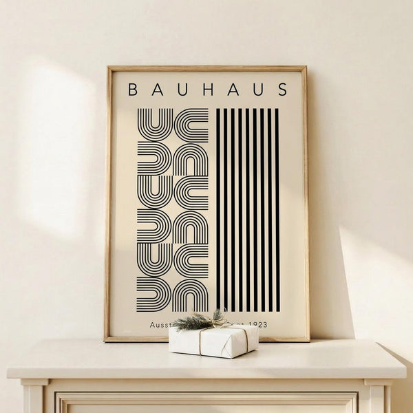 May include: Framed Bauhaus-style poster with geometric black line patterns and vertical stripes on cream background, displayed on light wood surface next to small wrapped gift with twine and greenery sprig against white wall