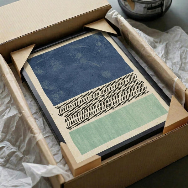 Abstract geometric print with horizontal bands of deep indigo blue at top, black and white herringbone chevron pattern in center, and sage green at bottom, all on cream background. The framed artwork sits in an open cardboard shipping box surrounded by white tissue paper, with wooden dowels visible nearby and part of a glass container in the background.