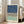 May include: Large framed abstract art print leaning against white wall next to beige sofa in bright living room. Artwork features three horizontal sections - textured navy blue at top, black and white herringbone pattern in middle, sage green at bottom. Light wood frame with white matting. Natural sunlight streams through tall windows onto hardwood floors.