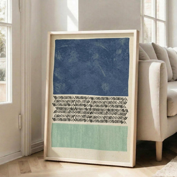 May include: Large framed abstract art print leaning against white wall next to beige sofa in bright living room. Artwork features three horizontal sections - textured navy blue at top, black and white herringbone pattern in middle, sage green at bottom. Light wood frame with white matting. Natural sunlight streams through tall windows onto hardwood floors.