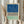 May include: Person holding a framed abstract art print with three horizontal sections - textured blue at top, black and white herringbone pattern in middle, and sage green at bottom. Light wood frame, neutral wall background, dried pampas grass visible to the left.